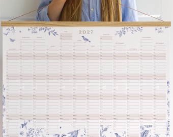 May include: A 2027 wall calendar featuring a vertical layout with twelve months, printed on white paper with blue floral and bird illustrations. The calendar is held by a light-colored wooden frame at the top and bottom, suspended by a thin brown cord.