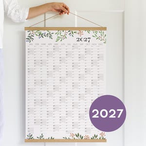 May include: A 2027 wall calendar printed on white paper with a grid layout for each month. The top and bottom edges are secured by light brown wooden magnetic poster hangers. The design features green botanical illustrations.