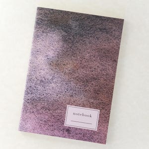 Op de afbeelding: Een rechthoekig notitieboek met een donkere, gevlekte cover in aquarel-stijl in tinten van dieppaars, zwart en gedempt bruin. Een wit rechthoekig label bevindt zich in de rechterbenedenhoek met het woord notebook in zwarte letters gedrukt.
