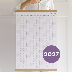 May include: A 2027 wall calendar printed on white paper with a grid layout for each month, featuring colourful headers for January through December. The calendar is held by a light wood frame at the top and bottom with a thin string for hanging.