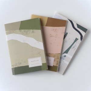 Puede incluir: Tres cuadernos con diseños abstractos de paisajes. Las cubiertas presentan colores apagados como verde, rosa y gris, con formas geométricas y la palabra "notebook" en uno. Los cuadernos están apilados en ángulo.