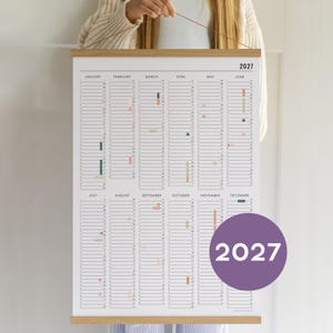 Peut inclure: Un calendrier mural 2027 avec une disposition verticale présentant les mois de janvier à décembre en colonnes. Le calendrier a un fond blanc avec du texte et des lignes noires, maintenu par un cadre en bois clair avec une ficelle.