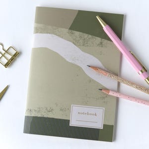 Puede incluir: Un cuaderno con un diseño abstracto en verde y blanco apagados, con la palabra "notebook" en una etiqueta. Un bolígrafo rosa y dos lápices, uno rosa y otro con un estampado de puntos, están junto al cuaderno. Un clip dorado está en la esquina superior izquierda.