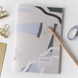 May include: A notebook with an abstract design in muted tones of beige, blue, and gray. A pencil rests on the cover, and the word "notebook" is printed on the front. Gold binder clips and a brass rectangle are visible.