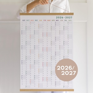 Peut inclure: Un calendrier mural pour 2026-2027 présentant une grille verticale avec les mois de septembre à août. Le calendrier est maintenu par un cadre en bois clair en haut et en bas. Une pastille beige affiche le texte 2026/2027 en blanc.