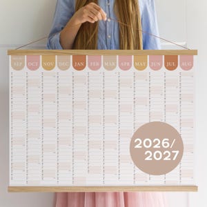 May include: A large, wall-mounted calendar for 2026 and 2027, featuring a minimalist design with a wooden frame. The calendar displays months from September to August, with a neutral color palette of beige, pink, and brown.
