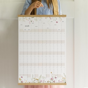 May include: A 2026 year-at-a-glance wall calendar with a floral design. The calendar is printed on white paper and features a grid layout with monthly columns and daily rows. The top and bottom edges are decorated with watercolor flowers and greenery. The calendar is held by wooden bars.
