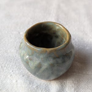 May include: A small, round ceramic vessel with a wide, open mouth and a thick, uneven rim. The exterior features a glossy, mottled glaze in shades of sage green, slate blue, and earthy brown, creating a textured, organic appearance throughout.