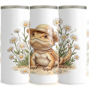May include: White tumbler with a cartoon lizard wearing a straw hat, surrounded by daisies. The lizard is tan and cream-coloured, with a friendly expression. The daisies have white petals and yellow centres. A charming and whimsical design.