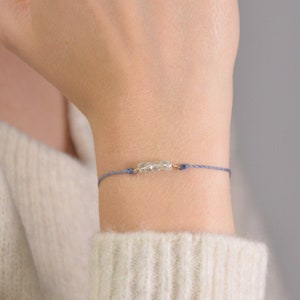 Puede incluir: Un delicado brazalete de cuerda azul con pequeños cristales transparentes y una cuenta dorada.