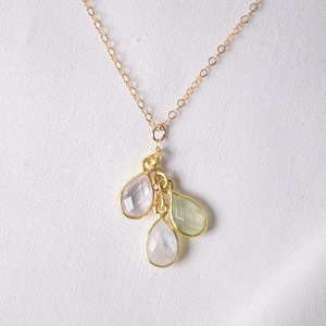 Puede incluir: Un collar de cadena dorada con un colgante que presenta tres piedras preciosas en forma de lágrima en tonos de blanco, rosa claro y verde.