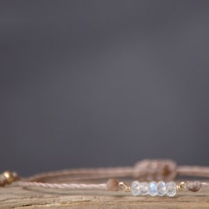 Op de afbeelding: Een delicate armband met een geknoopt beige koord en een rij heldere, iriserende kralen. De armband is geknoopt en heeft gouden accenten.