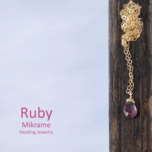 Puede incluir: Un collar de cadena dorada con un colgante de rubí rosa. El collar se muestra en un trozo de madera. El texto "Ruby Mikrame Healing Jewelry" es visible debajo del collar.