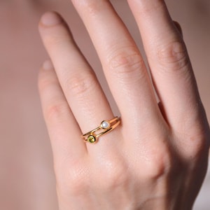 Puede incluir: Un anillo de oro con una pequeña piedra preciosa verde y una perla blanca. El anillo se lleva en el dedo anular de una mano.