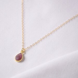Könnte beinhalten: Eine zarte goldene Halskette mit einem tropfenförmigen Anhänger. Der Anhänger hat einen rosa Edelstein, der in einer goldenen Lünette gefasst ist. Die Halskette ist fein und der Hintergrund ist ein weißer Stoff.