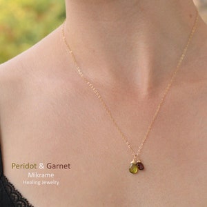 Könnte beinhalten: Eine goldene Kettenkette mit einem grünen Peridot- und einem roten Granatanhänger. Die Kette wird von einer Person getragen. Der Text "Peridot & Garnet Mikrame Healing Jewelry" befindet sich am unteren Rand des Bildes.