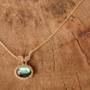 May include: A necklace with a light blue labradorite gemstone pendant, woven with macrame cord. The necklace is on a wooden surface.