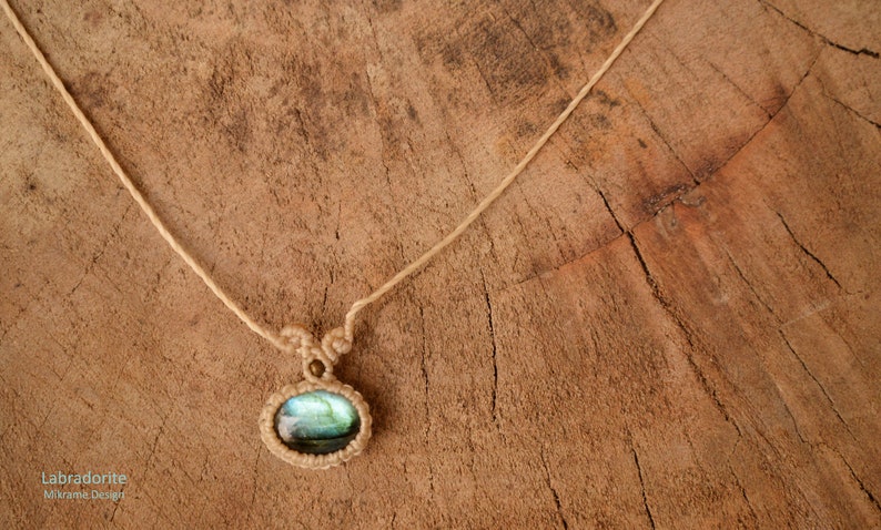 May include: A necklace with a light blue labradorite gemstone pendant, woven with macrame cord. The necklace is on a wooden surface.