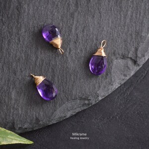 May include: Three amethyst gemstone pendants wrapped in gold wire. The pendants are on a dark grey slate surface. Mikrame Healing Jewelry is written in white text on the surface.