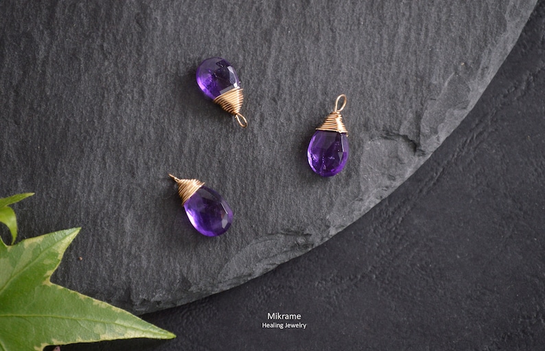 May include: Three amethyst gemstone pendants wrapped in gold wire. The pendants are on a dark grey slate surface. Mikrame Healing Jewelry is written in white text on the surface.