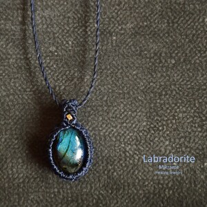 May include: A dark blue macrame necklace with a labradorite gemstone pendant. The pendant is oval-shaped and has a blue, green, and black iridescent sheen. The text "Labradorite Mikrame Healing Jewelry" is written below the pendant.