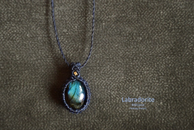 May include: A dark blue macrame necklace with a labradorite gemstone pendant. The pendant is oval-shaped and has a blue, green, and black iridescent sheen. The text "Labradorite Mikrame Healing Jewelry" is written below the pendant.
