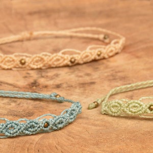 Puede incluir: Tres pulseras de macramé con un delicado patrón de ondas. Cada pulsera está hecha con un cordón de color diferente: azul claro, verde claro y crema. Las pulseras están adornadas con pequeñas cuentas doradas.