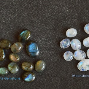 May include: A collection of Labradorite gemstones and Moonstone gemstones. The Labradorite gemstones are dark grey with blue and green iridescent flashes. The Moonstone gemstones are white with a blue iridescent sheen.  Labradorite Gemstone and Moonstone Gemstone.