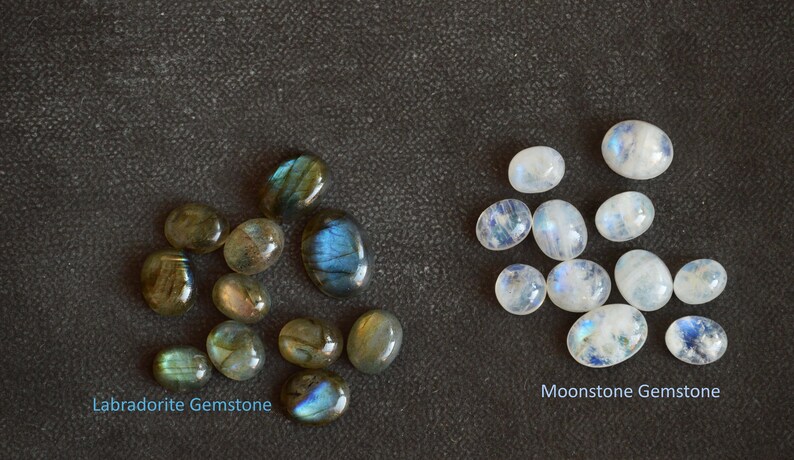 May include: A collection of Labradorite gemstones and Moonstone gemstones. The Labradorite gemstones are dark grey with blue and green iridescent flashes. The Moonstone gemstones are white with a blue iridescent sheen.  Labradorite Gemstone and Moonstone Gemstone.