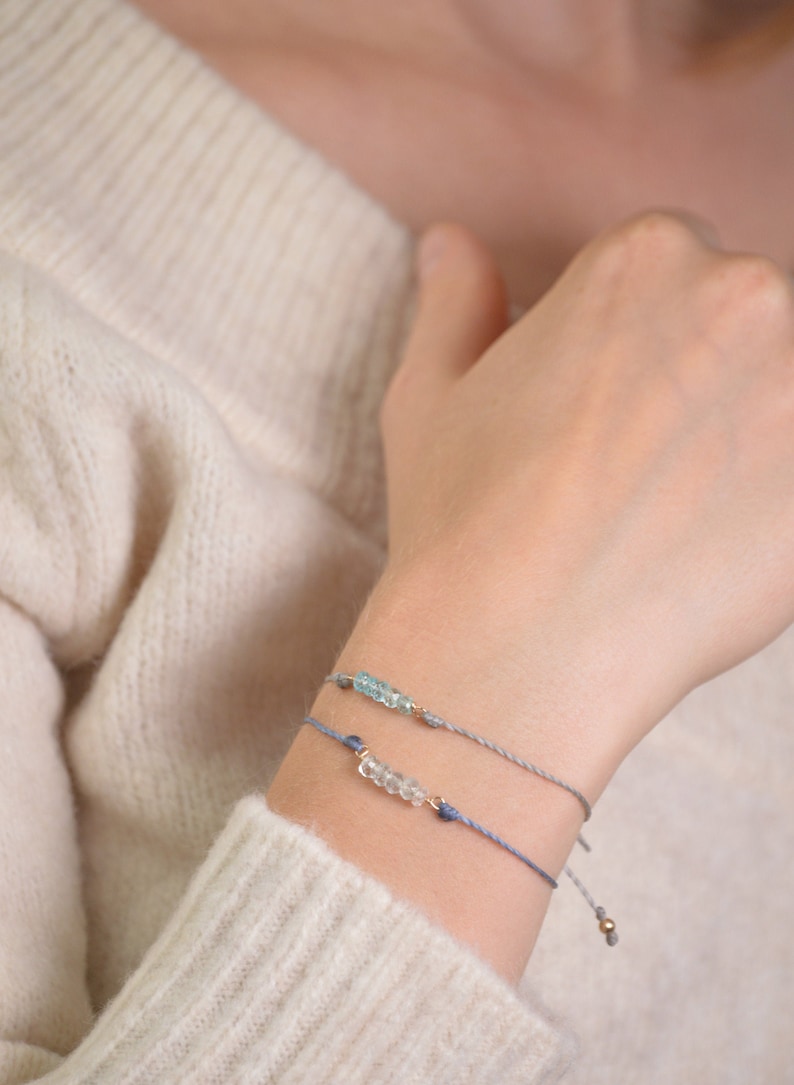 May include: Two delicate bracelets with light blue and clear gemstone beads on a light gray sweater.