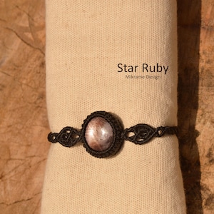 Puede incluir: Un brazalete de macramé negro con una piedra preciosa de rubí estrellado. El brazalete está sobre un fondo de tela beige. El texto "Star Ruby" y "Mikrame Design" es visible en la tela.