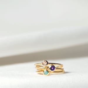 Puede incluir: Cuatro anillos de oro con piedras preciosas de diferentes colores. Los anillos están apilados uno encima del otro. Las piedras preciosas son de color rosa claro, azul claro, morado y transparente.