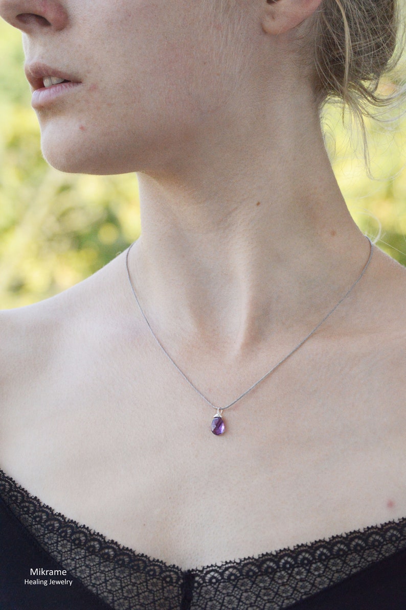 May include: A silver chain necklace with a small, purple gemstone pendant. The necklace is worn by a person with a black lace top.