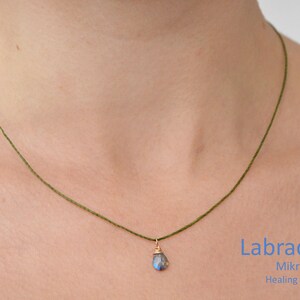 May include: A green cord necklace with a small, teardrop-shaped labradorite gemstone pendant. The pendant is set in gold and hangs from the cord. The text "Labradorite", "Mikrame", and "Healing Jewelry" is visible below the necklace.