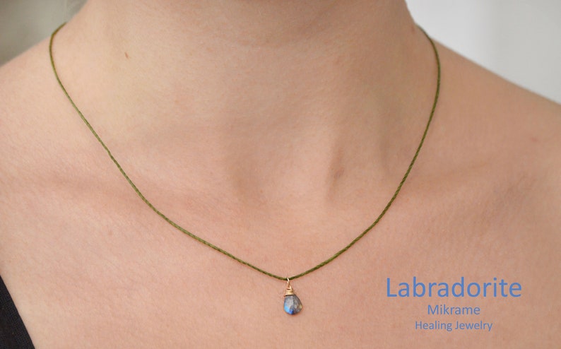 May include: A green cord necklace with a small, teardrop-shaped labradorite gemstone pendant. The pendant is set in gold and hangs from the cord. The text "Labradorite", "Mikrame", and "Healing Jewelry" is visible below the necklace.