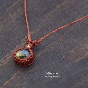 May include: A brown cord necklace with a round, green and gold labradorite gemstone pendant. The pendant is wrapped in brown cord and has a gold accent.