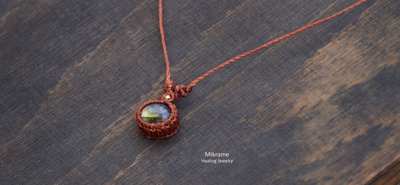 May include: A brown cord necklace with a round, green and gold labradorite gemstone pendant. The pendant is wrapped in brown cord and has a gold accent.