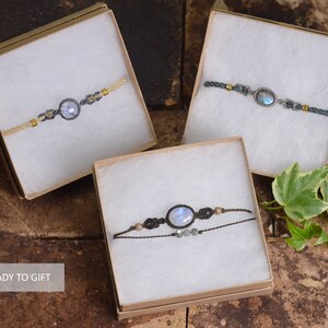 May include: Three gift boxes with white interior and brown exterior, each containing a different moonstone bracelet. The bracelets are made with brown cord and feature a moonstone gemstone. The boxes are sitting on a brown textured surface.  READY TO GIFT.