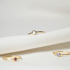 Puede incluir: Tres anillos de oro con piedras preciosas de diferentes colores. El anillo superior tiene una piedra blanca, el anillo del medio tiene una piedra morada y el anillo inferior tiene una piedra rosa.