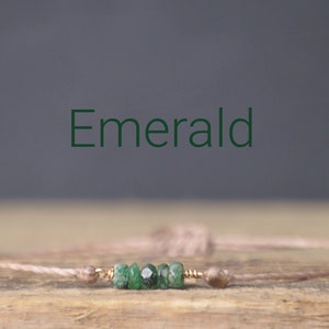 May include: A delicate bracelet with a light brown cord and a cluster of small, green emerald stones. The word "Emerald" is written in green text above the bracelet.