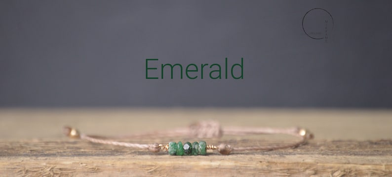 May include: A delicate bracelet with a light brown cord and a cluster of small, green emerald stones. The word "Emerald" is written in green text above the bracelet.
