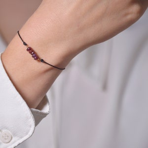 Puede incluir: Un brazalete sencillo con un cordón negro y una fila de pequeñas piedras preciosas de color rojo oscuro.