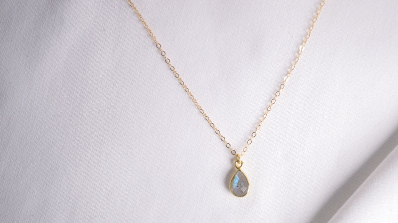 May include: A delicate gold chain necklace with a teardrop-shaped pendant featuring a clear iridescent gemstone.