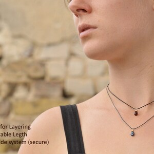 May include: Two delicate necklaces with small gemstone pendants. The top necklace has a red gemstone and the bottom necklace has a blue gemstone. Both necklaces are on a black cord and have a sliding clasp.