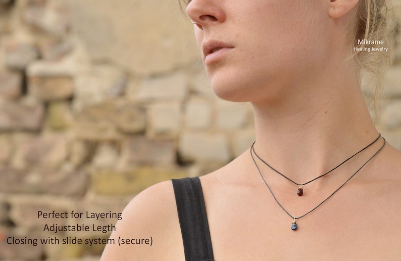 May include: Two delicate necklaces with small gemstone pendants. The top necklace has a red gemstone and the bottom necklace has a blue gemstone. Both necklaces are on a black cord and have a sliding clasp.