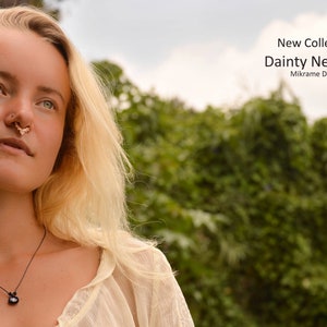 May include: A woman with blonde hair wearing a white shirt and a black necklace with a dark gemstone pendant. The necklace is a delicate, simple design. The woman is looking up and to the right. The background is blurred and shows green foliage.