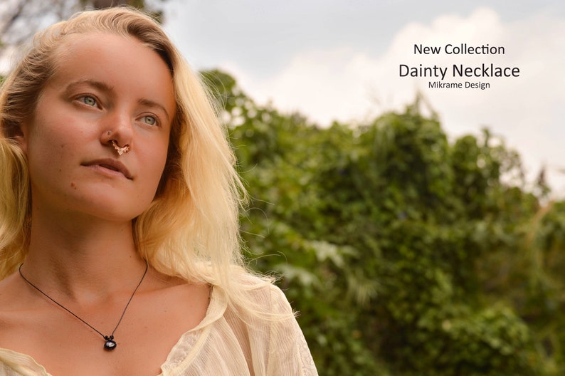 May include: A woman with blonde hair wearing a white shirt and a black necklace with a dark gemstone pendant. The necklace is a delicate, simple design. The woman is looking up and to the right. The background is blurred and shows green foliage.