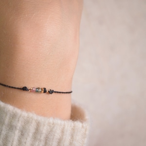 May include: A delicate black string bracelet with small, colorful beads. The beads are a mix of pink, green, and black.