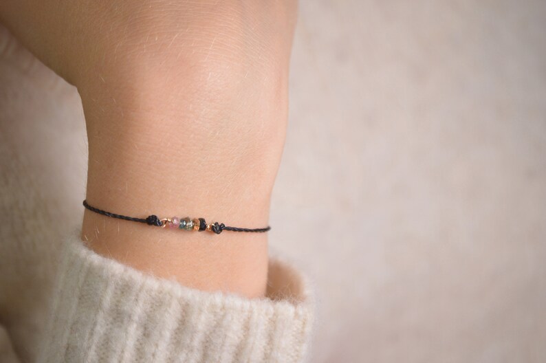 May include: A delicate black string bracelet with small, colorful beads. The beads are a mix of pink, green, and black.