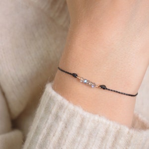 May include: A delicate black string bracelet with a small cluster of iridescent crystals and gold accents.
